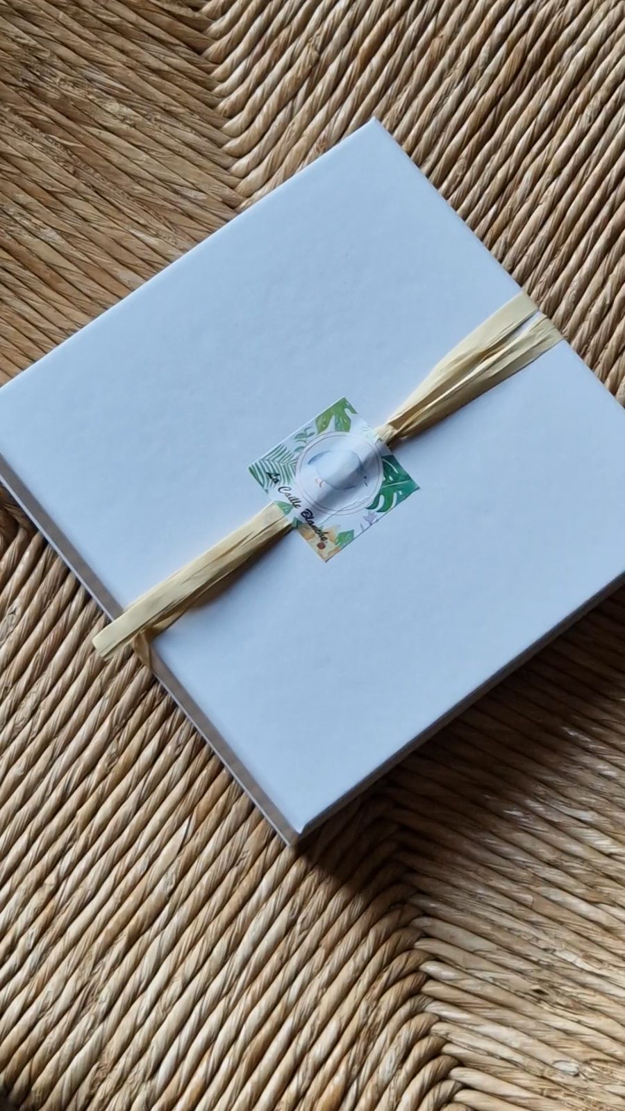 Coffret I — Le goût de La Caille Blanche, fermé, noué de raphia avec son autocollant, posé sur un plateau tressé