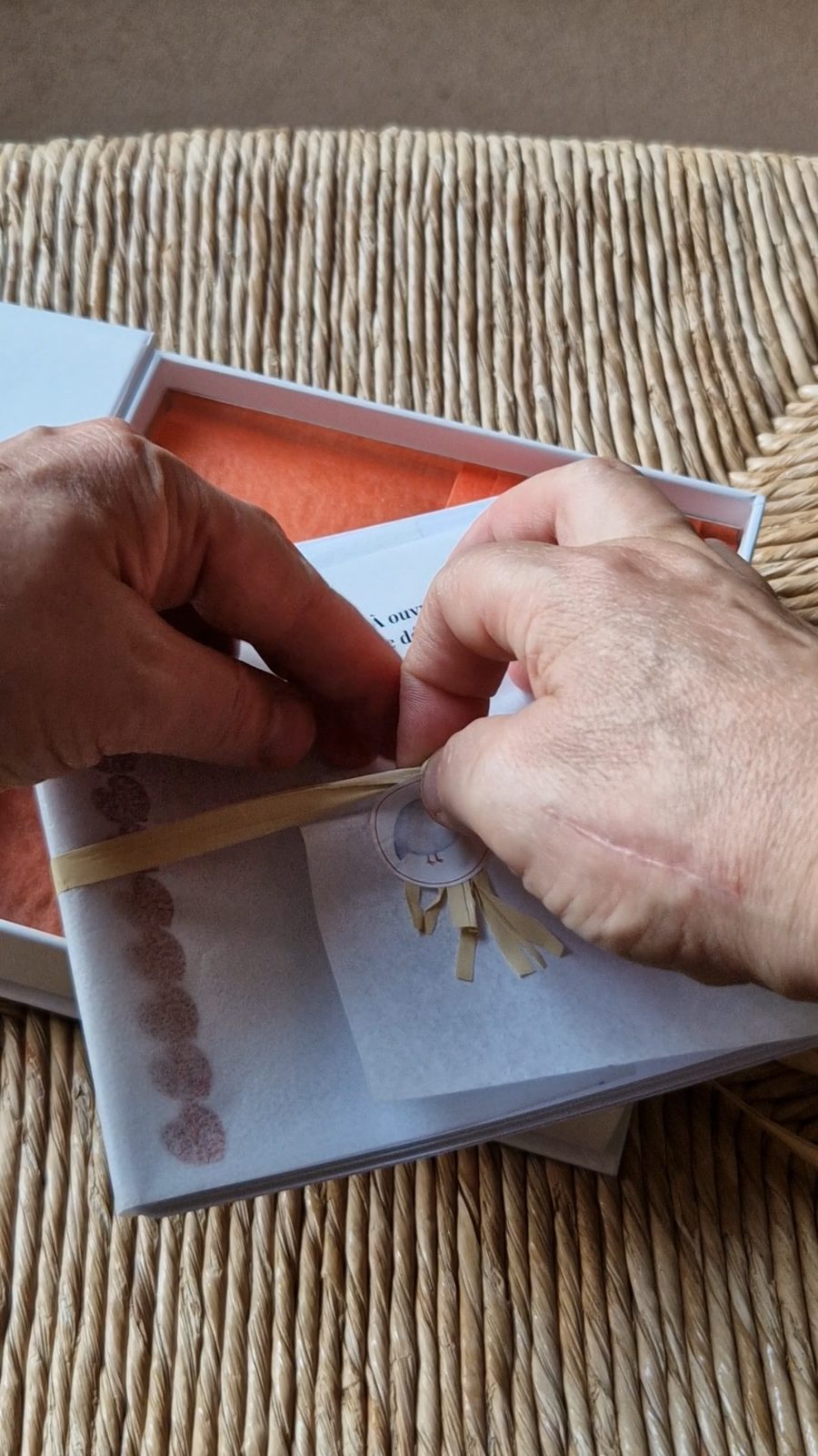 Mains dénouant le raphia du coffret I — Le goût, papier de soie orange dans la boîte ouverte